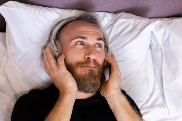 时髦快乐的白人男子戴着耳机躺在床上听他最喜欢的音乐 独自休息 跳舞科技家伙舒适