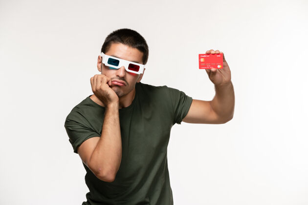 肖像正面图身着绿色t恤 戴着d墨镜 手持红色银行卡的年轻男子在白墙上拍摄电影《孤独电影院》电影院年轻男性男人