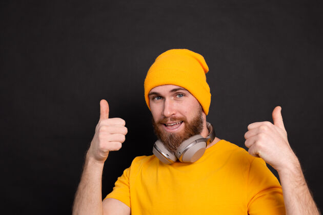 男性帅气的大胡子时髦男人黄色帽子t恤 黑色背景上有独立的耳机休闲手势手