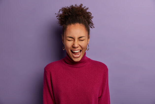 大笑快乐的黑皮肤女孩忍不住笑 眯着眼睛看笑话 傻笑 看一些可爱或有趣的东西 穿紫红色高领毛衣 孤立在紫色的墙上 有积极的一天肖像非洲室内