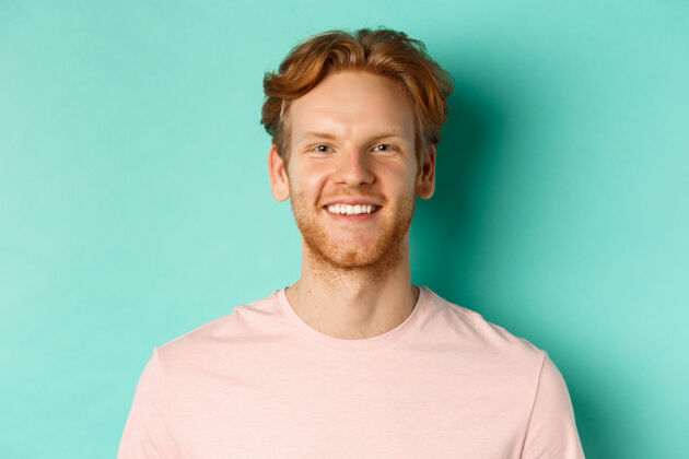 人特写镜头：红发胡子的家伙穿着粉色t恤 微笑着 牙齿洁白 看着相机 站在绿松石色的背景上脸男孩头发
