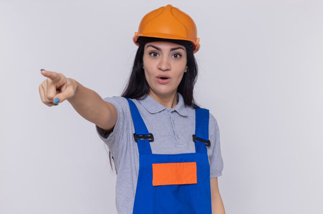 年轻人一位身穿建筑制服 头戴安全帽的年轻女建筑工人惊讶地用食指指着站在白墙上的东西目录建筑站
