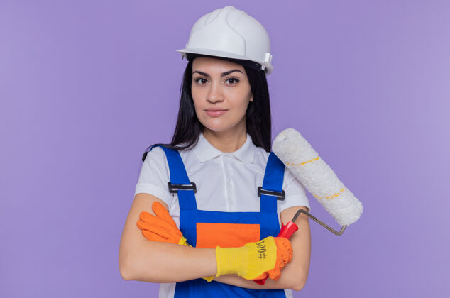 建筑工人身穿施工制服 头戴安全帽 头戴橡胶手套 手拿油漆辊 神情严肃自信地站在紫色的墙上 看着前方滚轴头盔橡胶
