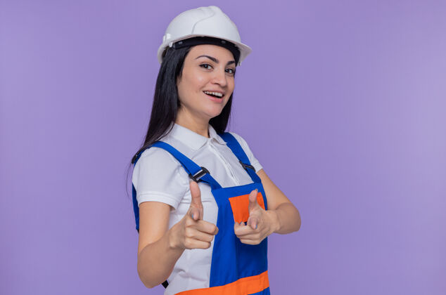 安全身穿施工制服 头戴安全帽的年轻建筑工人站在紫色的墙上 用食指指着前面 愉快地微笑着手指指点欢呼