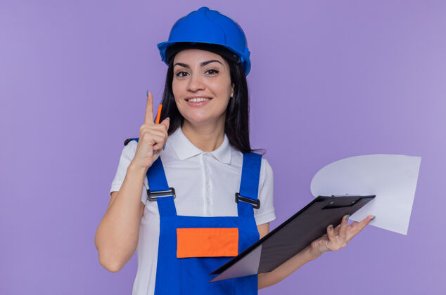 剪贴板身穿建筑制服 头戴安全帽 拿着写字板和铅笔的年轻建筑妇女站在紫色的墙上 面带微笑地看着前方微笑建筑工建筑