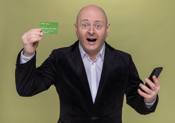 快乐穿着西装的中年秃头男人站在绿色的墙壁上展示信用卡的惊喜和快乐秀惊喜男人