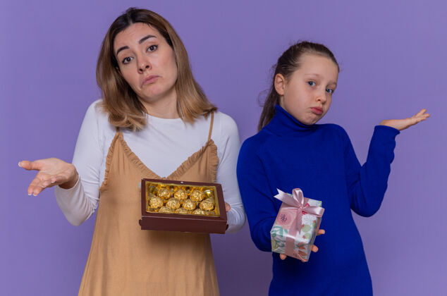 女人爱心妈妈拿着一盒巧克力 女儿拿着礼物 神情迷茫地举起双臂 站在紫色的墙上庆祝国际妇女节站着女儿困惑