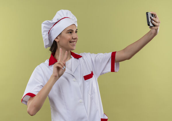 女孩身着厨师制服的年轻高加索厨师长微笑着看着电话 在绿色的空白处摆出胜利手势胜利标志年轻