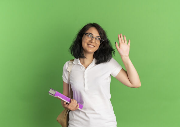 微笑年轻笑容可掬的白种女学生戴着眼镜 背着书包 举手看着旁边拿着书的绿色和复印空间壁板复制年轻