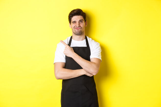 厨师不高兴的咖啡师穿着黑色围裙 指着左上角的手指 做着怀疑的表情 不喜欢什么东西 站在黄色的背景下成人站立秀