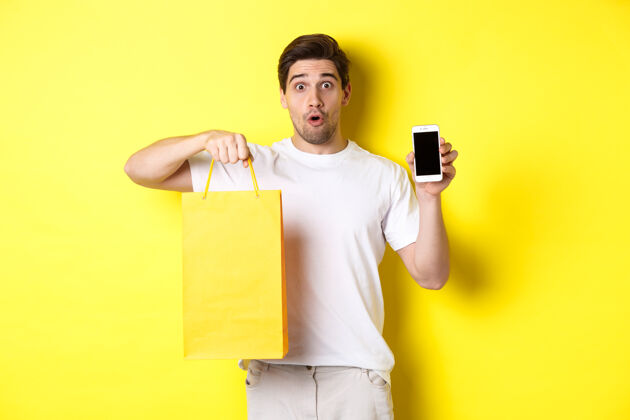 男人惊讶的男人拿着购物袋 展示智能手机屏幕 手机银行概念和应用成就 黄色背景手机黄色男人