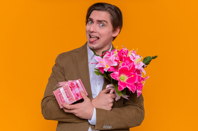 举行风趣的年轻人拿着礼物和花束望着前面伸出的舌头 准备站在橙色的墙上祝贺国际妇女节搞笑男人花束