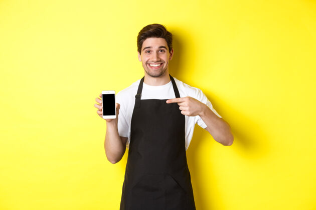 工作室穿着黑色围裙的帅哥咖啡师指着手机屏幕 微笑着展示应用程序 站在黄色背景上男士围裙男士