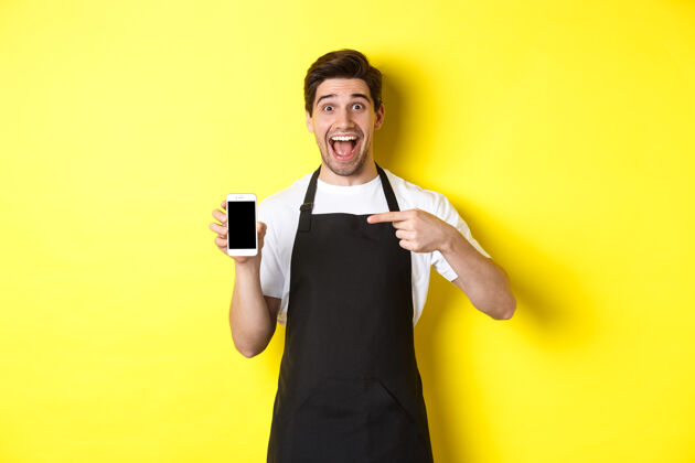 工作室穿着黑色围裙的帅哥咖啡师指着手机屏幕 微笑着展示应用程序 站在黄色背景上时尚年轻帅哥