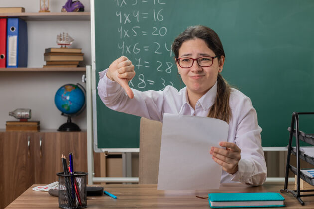 桌子戴着眼镜的年轻女教师坐在课桌旁 空白的书页看起来困惑和不高兴 在教室的黑板前竖起大拇指检查作业眼镜检查年轻
