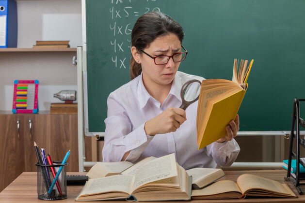 玻璃戴眼镜的年轻女教师坐在教室黑板前的课桌旁 用放大镜看书 感到困惑和不快老师戴着年轻
