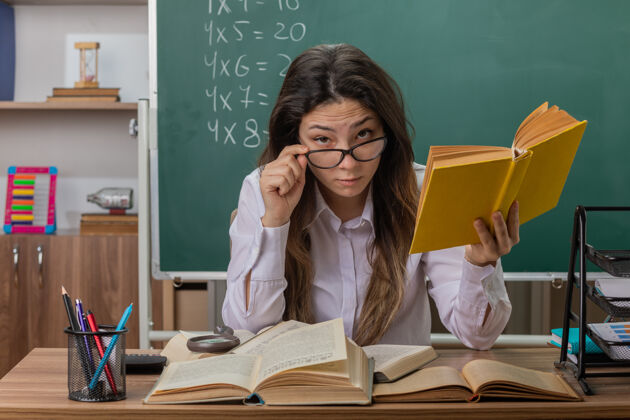 前面年轻的女老师戴着眼镜 专心地坐在教室黑板前的课桌旁看书老师坐着戴着