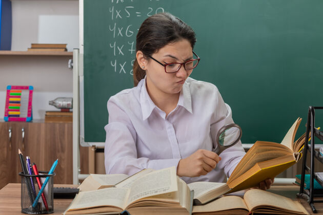 玻璃戴眼镜的年轻女教师坐在教室黑板前的课桌旁 用放大镜看书 感到困惑和不快穿过老师前面