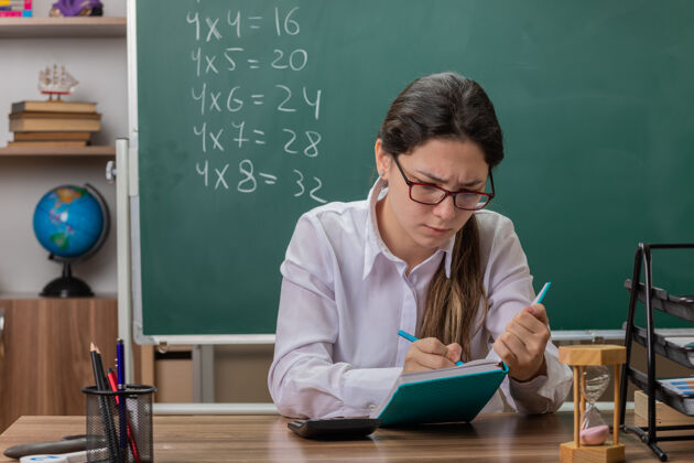 课戴眼镜的年轻女老师在备课笔记本上写东西 坐在教室黑板前的课桌旁 看起来很自信东西笔记本教室