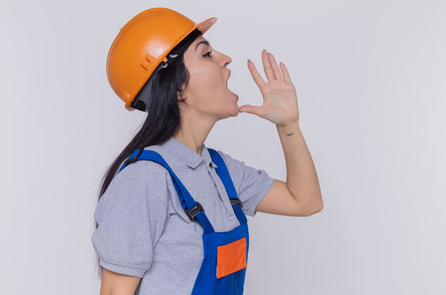 制服身穿施工制服 头戴安全帽的年轻建筑工人站在白色的墙壁上大声喊叫或叫着别人站立女人呼喊