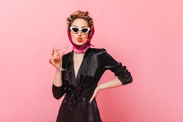 欢呼戴墨镜的女士在粉红色的墙上摆姿势 拿着马提尼杯 吃着橄榄黑发室内优雅
