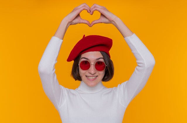 头快乐的年轻女子 短发 白色高领毛衣 头戴贝雷帽 戴着红色太阳镜 微笑着 手指举过头顶 站在橙色的墙上 做着心形手势太阳镜穿头发