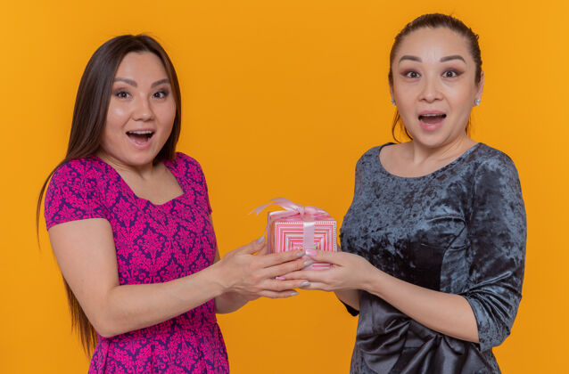 亚洲两位欢庆国际妇女节的亚洲妇女手拿礼物站在橙色的墙上 脸上洋溢着笑容现在举办庆祝