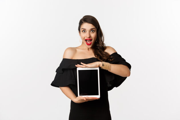 女性时尚和购物理念令人惊叹的年轻女子在平板电脑屏幕上展示在线网站促销活动 看起来兴奋的相机 站在黑色礼服 白色背景美丽年轻女性