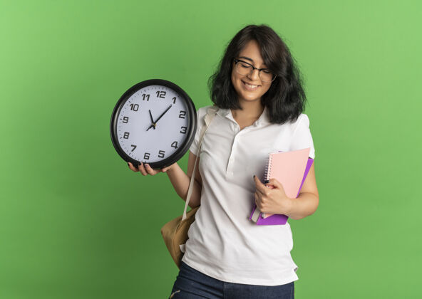 时钟年轻的笑容可掬的白人女学生戴着眼镜 背着书包 拿着时钟和笔记本放在绿色的空白处背部眼镜复制