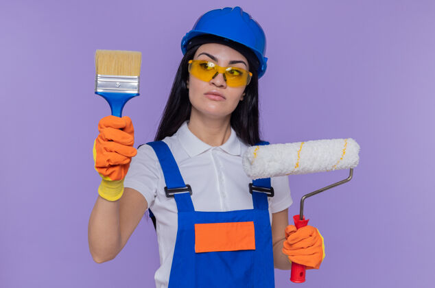 戴着身穿施工制服 戴着安全帽 戴着橡胶手套 拿着油漆辊和刷子的年轻建筑工人站在紫色的墙上严肃地看着它拿着滚轴女人