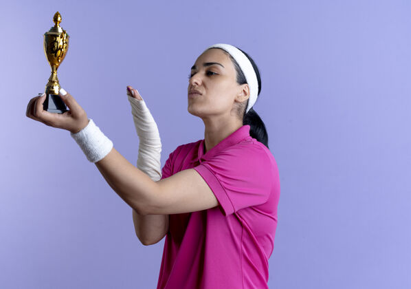 运动年轻自信的高加索运动女性戴着头带和腕带拿着并指着紫色的冠军杯穿女子腕带