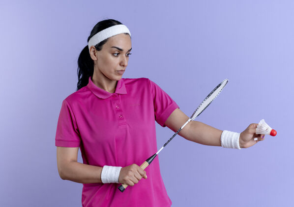 太空年轻严肃的白人运动女性戴着头带和腕带拿着和看着羽毛球拿着球拍在紫色的复制空间运动持有复制