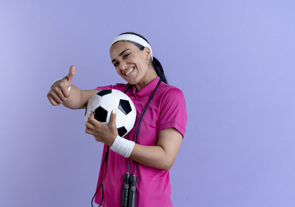 女人年轻微笑的白人运动女性戴着头带和腕带 脖子上系着跳绳 拿着球大拇指朝上放在紫色的复制空间上拇指复制腕带
