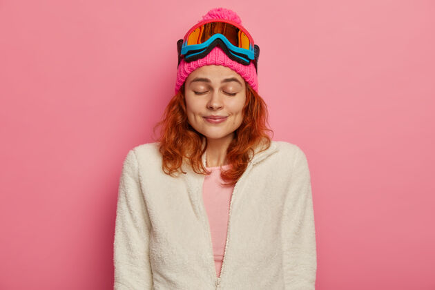 室内迷人的红发女人戴着暖和的帽子 滑雪镜和白色套头衫跳伞者请表达