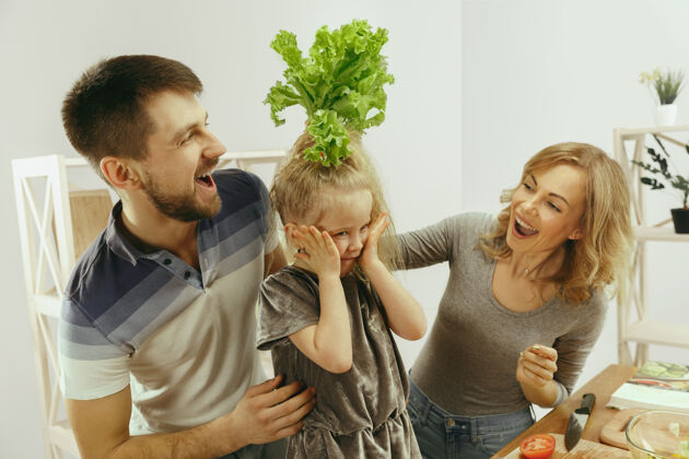 食物可爱的小女孩和她美丽的父母在家的厨房里切蔬菜 一边微笑着做沙拉美丽厨房积极
