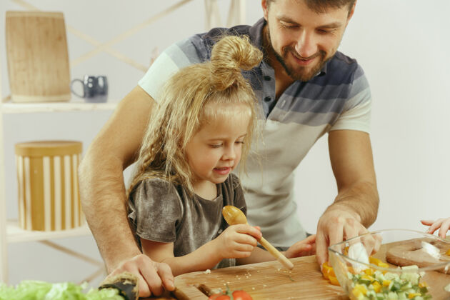 小可爱的小女孩和她美丽的父母在家的厨房里切蔬菜 一边微笑着做沙拉男性爸爸一起