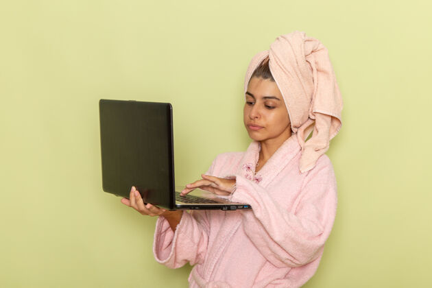 年轻女性前视图穿着粉红色浴袍的年轻女性在绿色表面上使用笔记本电脑漂亮前面笔记本电脑