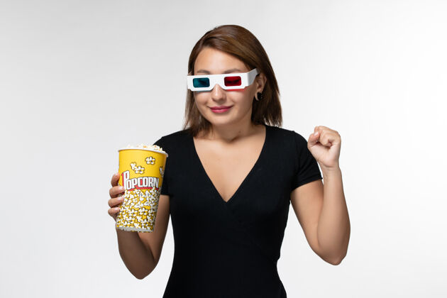 年轻的女性正面图：戴着d型太阳镜拿着爆米花包的年轻女性 在白色的表面上微笑电影爆米花美丽