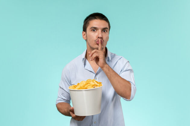 问正面图：年轻的男性拿着土豆片的篮子 要求在蓝色的表面上保持沉默电影院孤独年轻的男