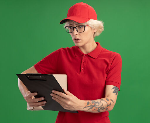 剪贴板年轻的送货员身穿红色制服 戴着帽子 戴着眼镜 手里拿着纸板箱 手里拿着剪贴板 焦急地站在绿色的墙上看着它制服立场担心