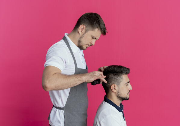 围裙穿着围裙的理发师在粉色墙上用满意客户的理发机理发满意男人顾客