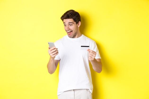 年轻男人惊讶地看着智能手机 网上购物 拿着信用卡 站在黄色背景上复制空间表情工作室站着