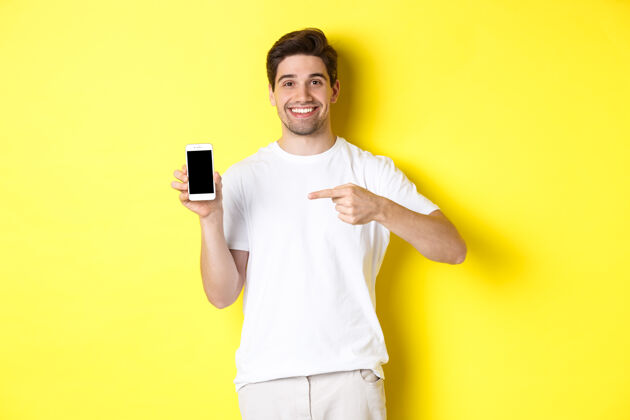 时尚一个漂亮的年轻人用手指着智能手机屏幕 展示一个应用程序 站在对面黄色模特自信