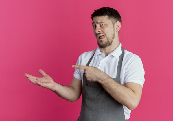 男人围裙上的理发师手拿着一个东西 用食指指着站在粉红色墙上的一边围裙东西指向