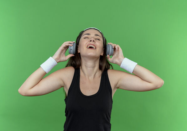 积极戴着耳机戴着头带的年轻健身女士站在绿色的墙上 快乐而积极地欣赏着她最喜爱的音乐头带喜爱音乐
