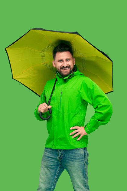 站立帅哥留着胡子 微笑着 拿着雨伞 看着前面孤立的绿色工作室 生动时尚夹克成人时尚