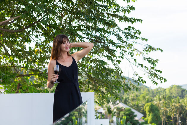时尚在热带阳台上 一个穿着黑色晚礼服 端着一杯葡萄酒的女人女性放松热带