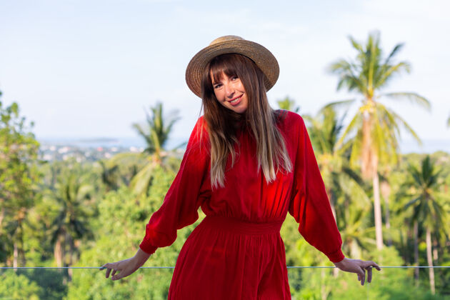 脱毛穿着红色夏装 戴着草帽 在阳台上享受热带海景和棕榈树的快乐女人时尚站立沙滩