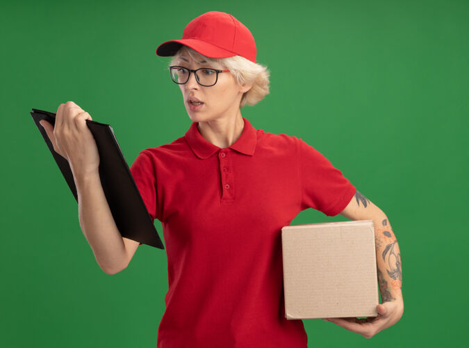制服年轻的送货员身穿红色制服 戴着帽子 戴着眼镜 拿着纸板箱 焦急地看着剪贴板 站在绿色的墙上戴着女人站着