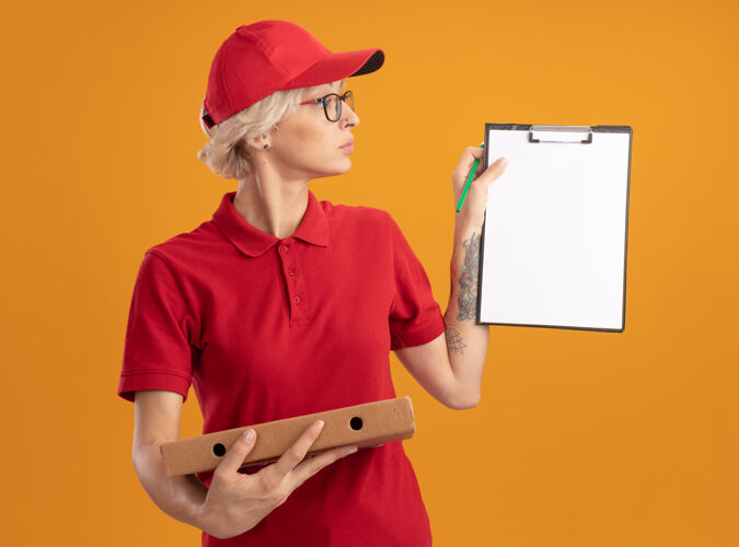 严肃身穿红色制服 戴着眼镜 拿着披萨盒的年轻送货员站在橙色的墙上严肃地看着披萨盒披萨女人穿
